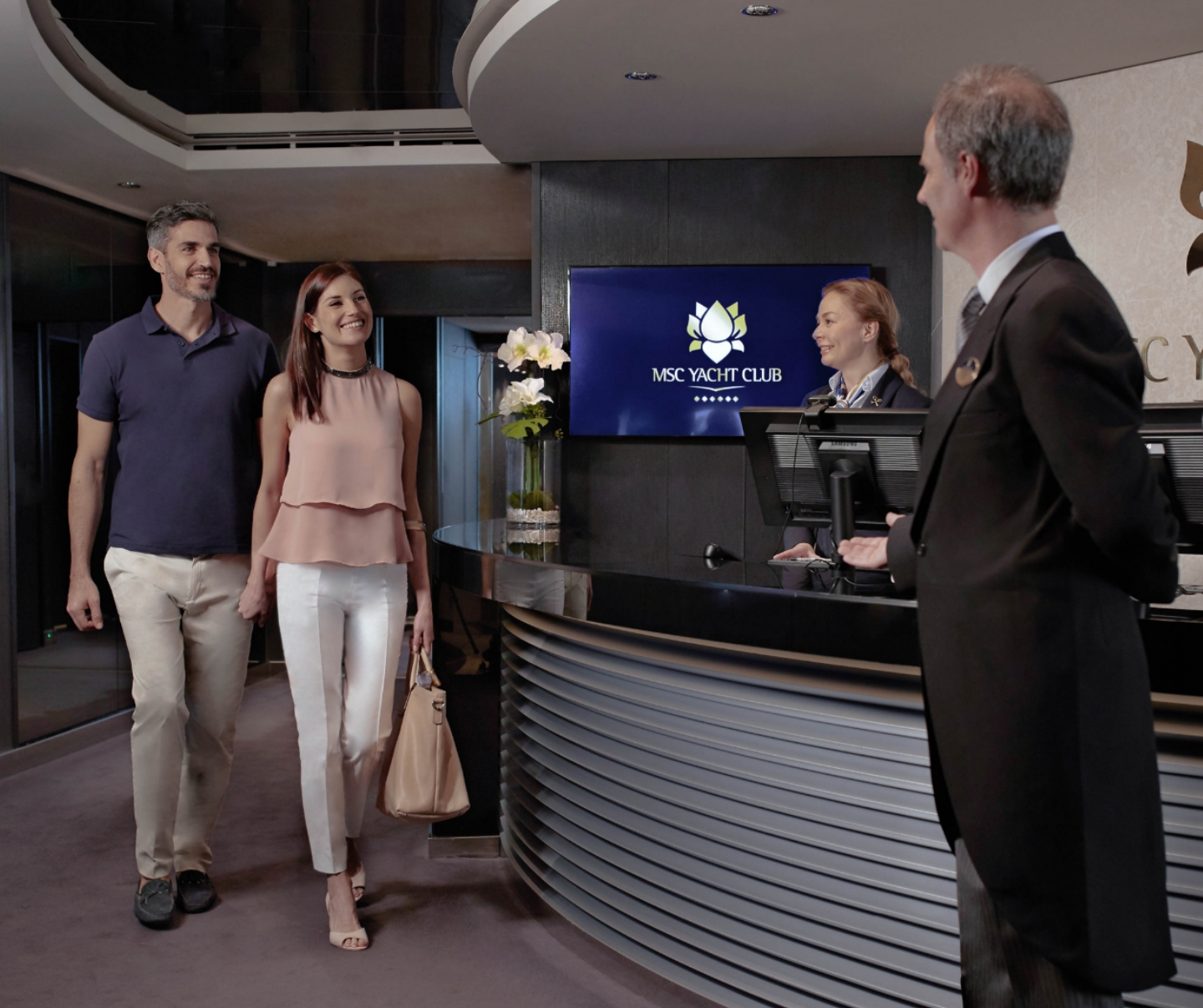 Young couple entering the Yacht Club's front lobby, where they enjoy the perks of luggage delivery and personalized service from the butler, catering to their cruise needs. 