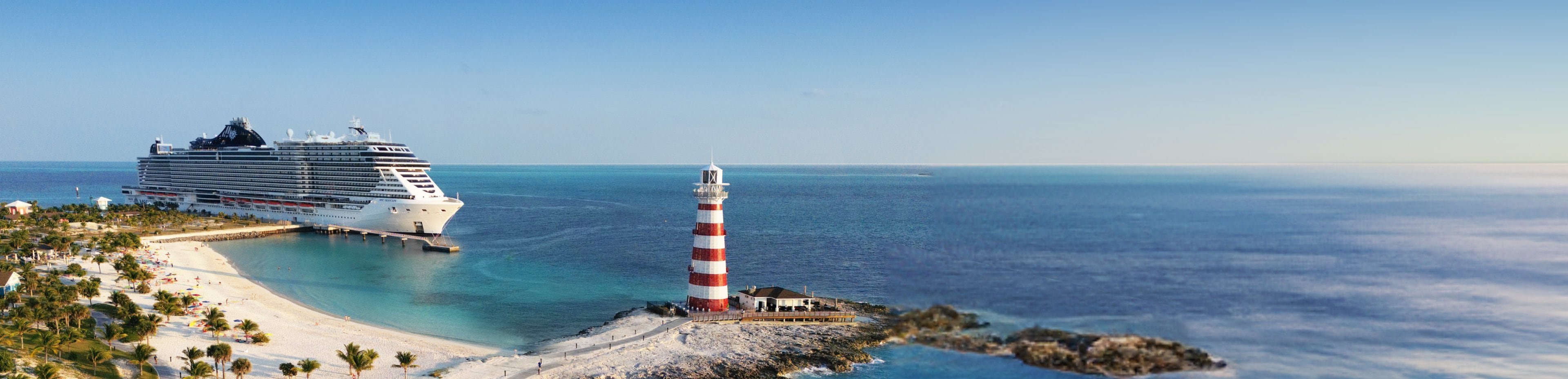 A beautiful shot of MSC Seascape docked at a Ocean Cays sandy beach, with a towering lighthouse in the background.