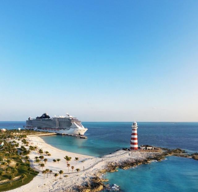 A beautiful shot of MSC Seascape docked at a Ocean Cays sandy beach, with a towering lighthouse in the background.