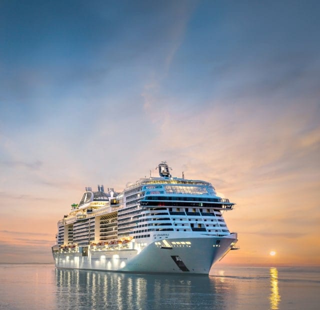 MSC Grandiosa sailing on an open sea at dusk with a setting sun in the background.